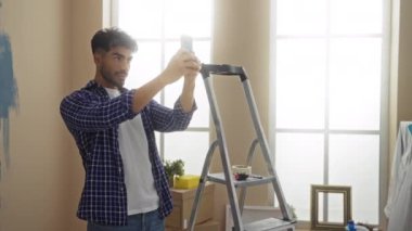 Sakallı genç adam yeni evini yenilerken selfie çekiyor. Etrafı boya malzemeleri, merdiven ve oturma odasındaki kutular ile çevrili..