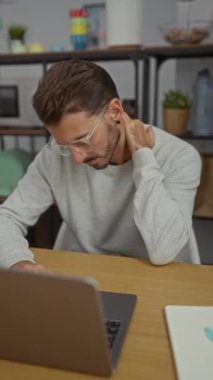 Ofisteki genç adam dizüstü bilgisayarla çalışırken boynunu tutuyor, bu da stres ya da yorgunluk anlamına geliyor..