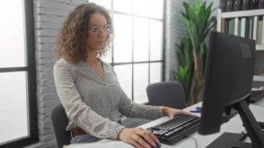 Modern işyerinde masa başında gülümseyen bir kadın bilgisayar, gözlük ve bitki ile doğal ışık ile profesyonel, sıradan bir atmosfer yaratıyor..