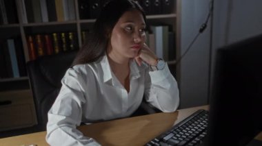 Ofiste çalışan bir kadın, bilgisayarla masasında otururken yorgun görünüyor, arka planda kitaplıklar, loş ışıklandırma, beyaz gömlek, saat, esmer, yetişkin, kadın, ev içi, işyeri