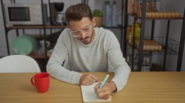 Gözlüklü genç adam bir ofiste bir masada oturuyor. Elinde defter, kalem ve kırmızı kupa var. Kollarını kavuştururken düşünceli görünüyor ve ileriye bakıyor..