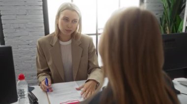Ofiste sarı saçlı bir kadın, modern iş yerindeki iş arkadaşlarıyla iş belgelerini tartışıyor. Masaüstü bilgisayarları ve en az dekor içeren bir ortam.