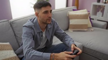 Oturma odasındaki rahat bir kanepede oynayan bir adam, elinde günlük giysiler giyen bir kontrolörle ekrana odaklanmış.