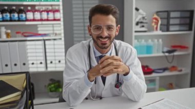 Hastane odasında tıbbi ekipman ve dosyalarla güvenle gülümseyen genç doktor karşılama kliniği ortamı yaratıyor..