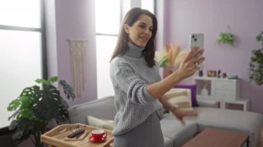 Gri kazaklı, şık oturma odasında selfie çeken bir kadın modern bir ev atmosferi sergiliyor..
