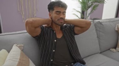 Kanepede uzanan genç bir adam. Elleri kapalı, rahat bir dairede. Bitkiler ve yastıklarla kurulmuş..