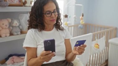Bebek eşyalarıyla dolu rahat bir kreş odasında telefon ve kredi kartı kullanan yetişkin bir kadın. İnternetten alışveriş ya da evde mali işlemleri gösteriyor..