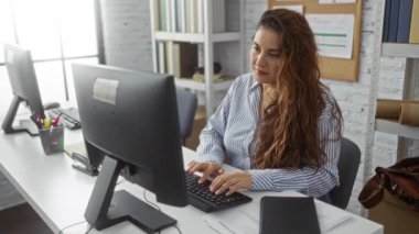 Ofiste bilgisayar ve belgelerle çalışan, odaklanmış ve profesyonel görünen bir kadın, çoklu görev becerilerini vurgulayan modern bir çalışma ortamı yakalıyor.