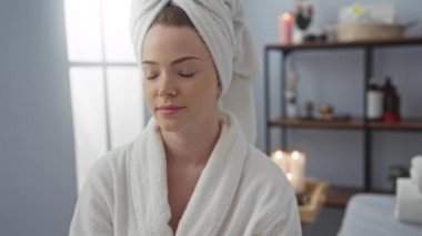 Kafasında havlu olan genç bir kadın spa salonunda beyaz bornoz giyerek dinleniyor.