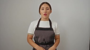 Önlüklü genç İspanyol kadın şaşkınlık ve şaşkınlık dolu bir yüzle duruyor, ağzı kapalı beyaz arka planda şüpheyle bakıyor.