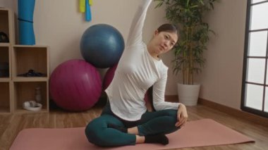 Spor salonundaki kadın egzersiz topları ve bitkilerle paspasın üzerinde geriniyor, huzurlu bir duruş sergiliyor ve sakin bir oda atmosferine odaklanıyor..