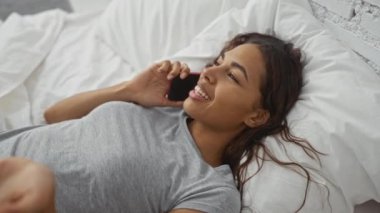 Yatak odasında rahat bir şekilde uzanmış, gülümseyen ve rahatlamış bir şekilde telefonla konuşan genç bir kadın.