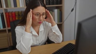 Ofis işyerindeki İspanyol kadın kafasını elinde tutarken ve bilgisayar ekranına bakarken stresli ve hayal kırıklığına uğramış görünüyor..