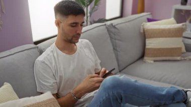 Evdeki akıllı telefonu kullanan genç adam oturma odasındaki kanepede dinleniyor gündelik kıyafetlerle düşünceli ve şık görünüyor, sakin ve modern bir atmosfer yaratıyor..
