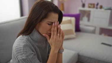 Oturma odasındaki kanepede oturan, elleri yüzü kapalı, gri bir kazak giyen, duygularını gösteren, güzel, rahat atmosferli esmer, yetişkin bir kadın..