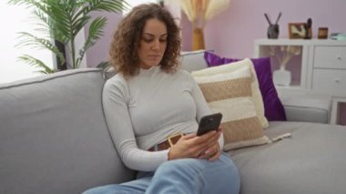 Rahat bir oturma odasında rahat bir kanepede dinlenen, yorgun bir ifadeyle akıllı telefonuna bakan, yastıklar ve modern dekorlarla çevrili boyun ağrısını öneren bir kadın..