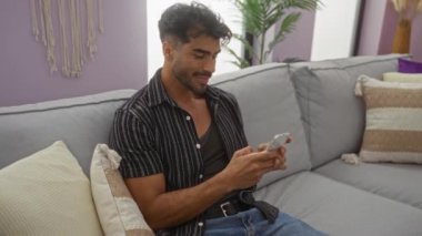 Sakallı genç adam rahat oturma odasındaki kanepede akıllı telefon kullanıyor. Canlı apartman ortamında keyif veriyor..