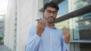 Kentsel bir sokakta duran genç adam, arka planda modern şehir binaları olan, gözlüklü ve mavi gömlekli bir akıllı telefon kullanarak sesli mesaj kaydediyor..