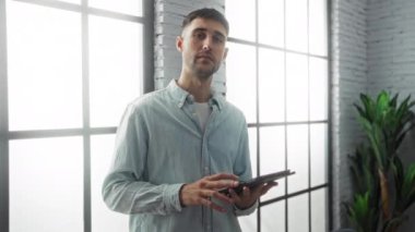 Geniş pencereli modern bir ofiste tablet kullanan genç adam profesyonel ve kendinden emin bir atmosfer sergiliyor..