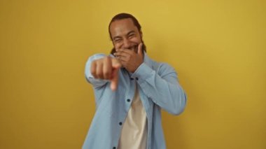 Sakallı genç adam izole edilmiş sarı arka planda gülüyor mavi gömlekli ve şakacı bir şekilde ileri doğru işaret ediyor..