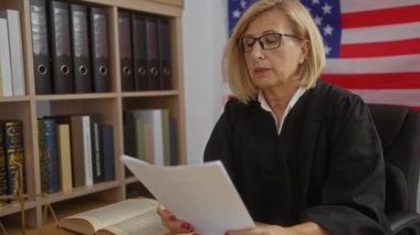 Kadın yargıç, arka planda kitap rafları ve bayraklarla Amerikan mahkemesinde belge okuyor.