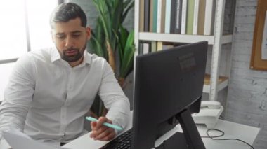 Ofiste çalışan genç bir adam masasında bilgisayar monitörüyle dokümanları inceliyor. Doğal güneş ışığıyla zenginleştirilmiş modern bir ev ortamında odaklanmış bir çalışma ortamı yaratıyor..