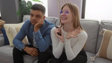 Evdeki kanepede oturan çift rahat bir şekilde oturma odasında oturuyor. Yastıklar ve doğal ışık pencerelerden içeri giriyor. Kadın gülümsüyor, erkek düşünceli görünüyor.