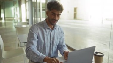 Modern bir ofiste dizüstü bilgisayarda çalışan sakallı genç adam kollarını kavuşturarak geniş pencerelerden içeri ışık sızan odaklanmış ve profesyonel bir atmosfer yaratıyor..