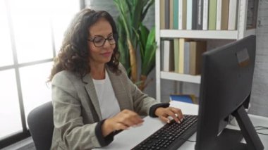 Modern ofiste bilgisayar ve tablet kullanan, birden fazla görev ve odaklanma sergileyen kitaplar ve bitkilerle çevrili bir kadın..