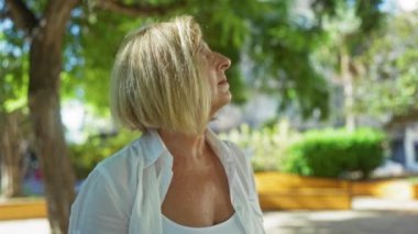 Şehir parkında dışarıda duran kısa sarı saçlı bir kadın yeşil ağaçlar ve güneş ışığının içinden geçtiği şehir ortamında düşünceli bir ifadeyle yukarı bakıyor..