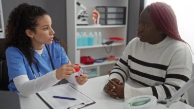 Mavi üniformalı kadın klinikteki Afrikalı Amerikalı kadına tıbbi prosedürü açıklıyor. Arka planda tıbbi malzemeler var..