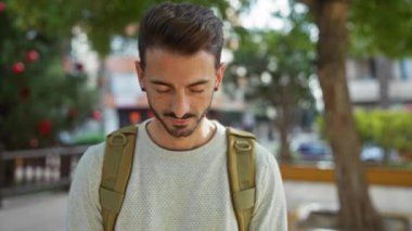 Şehir parkında sırtüstü sırtüstü sırtüstü sırtüstü sırtüstü gülümseyen genç bir adam, yakışıklı ve açık havada şehrin arka planına odaklanarak kendine güveni ve tarzını ifade ediyor..