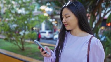 Şehir parkında şehir arka planında bulanık telefon kullanan, modern yaşam tarzını ve dışarıdaki bağlantılarını gösteren bir kadın..