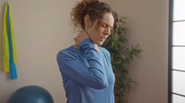 Mavi gömlekli, boynu ağrıyan, kapalı spor salonunda fizyoterapi bağlamında rahatsızlığını gösteren bir kadın..