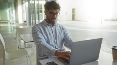 Ofiste dizüstü bilgisayarla çalışan, gözlüklü ve rahat gömlekli genç bir İspanyol, odak ve konsantrasyonu modern kapalı bir iş ortamında yansıtıyor..