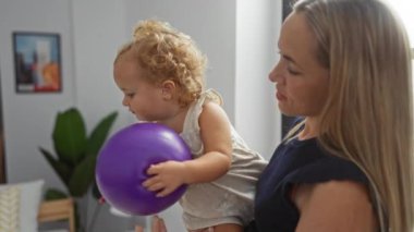 Mor topla küçük kızı yatak odasında tutan kadın, aile sevgisini sergiliyor, içeride iç mekan unsurlarıyla ev dekorasyonunu ön plana çıkarıyor..