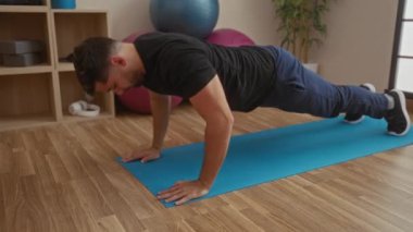 Genç adam mavi bir paspasın üzerinde egzersiz yapıyor, spor salonunda şınav çekiyor, spor aletleri ile çevrili, güç gösterisi ve atletik adanmışlık yapıyor..