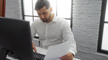 Beyaz gömlekli İspanyol bir adam modern bir ofis masasında, parlak, minimalist bir işyerinde bilgisayarı olan belgeleri inceliyor, odak ve profesyonelliği aktarıyor..