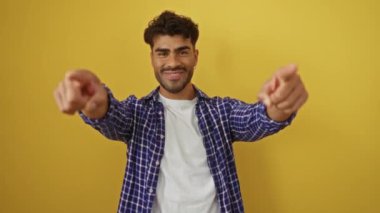 Hispanik bir adam canlı sarı bir arka plana karşı kollarını açmış gülümsüyor, günlük bir ekose gömlek ve beyaz tişört giyiyor, dostluk ve yakınlık taşıyor..