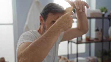 Orta yaşlı bir adam kaplıcada saçlarını tarıyor. Rafları ve doğal ışığı var. Huzurlu ve odaklanmış bir ortam yayıyor..