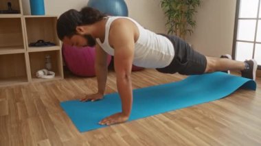 Sakallı genç adam, egzersiz topları ve ahşap zeminle çevrili, iyi aydınlatılmış bir spor salonunda, mavi bir paspasın üzerinde şınav çekiyor..