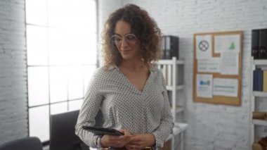 Modern ofis ortamında kameraya doğru uzanan kadın kıvırcık saçlı ve gözlüklü elinde tablet ve duvarlarda sergilenen belgelerle.