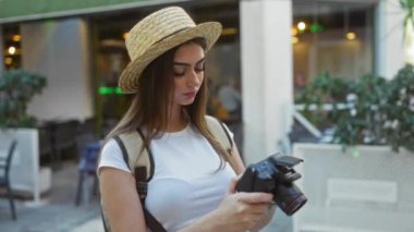 Şehir ortamında elinde hasır şapka ve beyaz gömlekle gezen, sırt çantalı, kumral saçlı, odaklanmış ve şık görünümlü genç bir kadın..