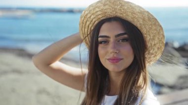 Deniz kenarındaki gezinti güvertesinde gülümseyen bir kadın parlak güneş ışığı altında hasır şapka takıyor ve okyanus zemininde canlı mavi su ve yaz atmosferinde neşeli ifadesini vurguluyor..