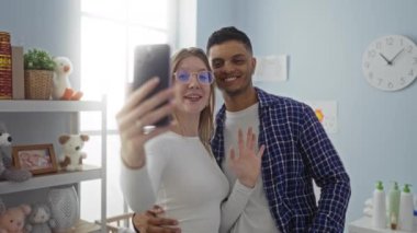 Parlak yatak odasında selfie çeken çift, ellerinde sevgi ve bağlılık gösteren kameralar, oyuncaklar ve dekorlarla çevrili ev ortamını güzelleştiriyor.