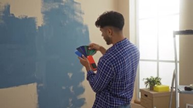 Yeni evin oturma odasında boya rengi seçen genç bir adam, duvarın yanında renk örnekleri tutan, pencereden içeri giren doğal ışık, iç dekorasyon projesi planlayan bir adam..