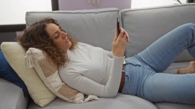 Kadın rahat bir oturma odasında kanepeye uzanmış, esniyor ve akıllı telefon kullanıyor. Evinde rahat bir atmosfer sergiliyor. Modern bir iç mekan..