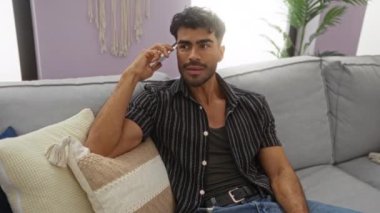 Oturma odasında telefonla konuşan İspanyol bir adam, gündelik kıyafetlerle kanepede oturuyor, pencereden süzülen parlak bir ışık, modern ev yaşam tarzını ve rahatlığını gösteriyor..