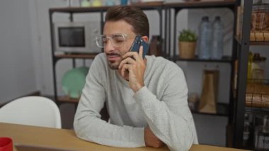Günümüz dekorasyonu ve ofis malzemeleriyle çevrili, gündelik kıyafetlerle telefonda konuşan genç adam, modern bir çalışma ortamı sergiliyor..