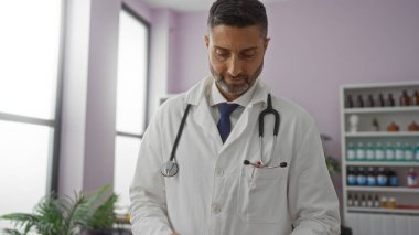 Modern klinikte steteskoplu beyaz önlüklü erkek doktor pencere kenarındaki hasta dosyasını inceliyor.